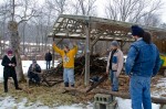 Creek Iverson, manager of the Brook Farm Project (in the center wearing the yellow sweater), passes over the saw to volunteer Zach Baker of NYC, so that he may teach the lumberjack song to the group for a sing a long. Photo by Dawna M. Cservak.